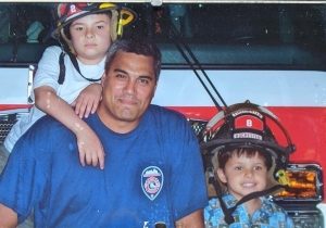FF Elvis Reyes sitting on the bumper of Truck 10 with young Reid Standing behind him leaning on his shoulder wearing a helmet and young Remmy sitting beside Elvis wearing a fire helmet. 